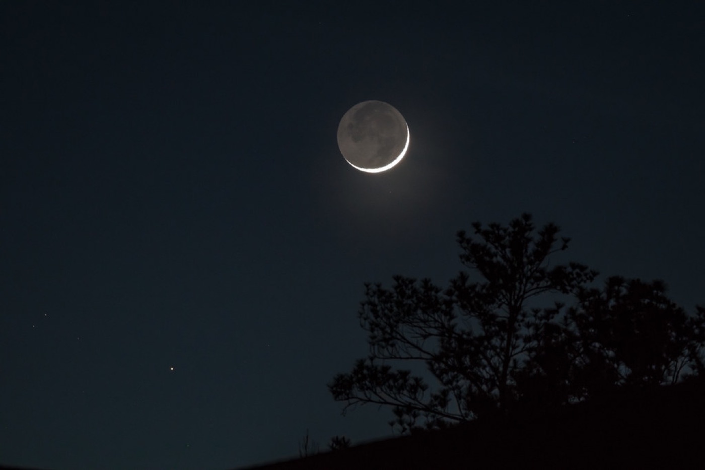 A thin crescent moon above a calm dark horizon with a single candle in the foreground.