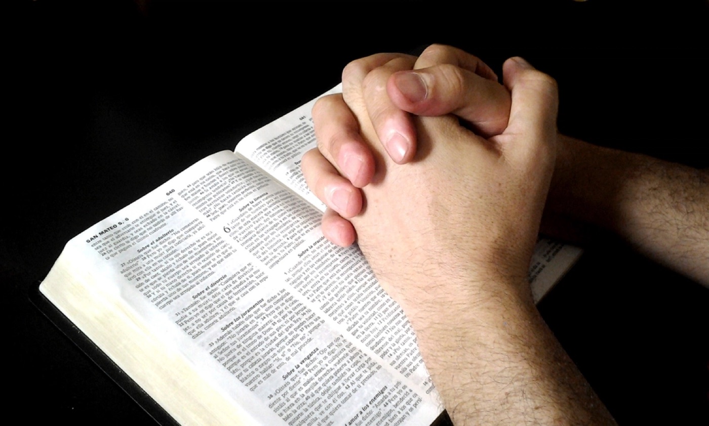 Hands folded in prayer with soft golden light falling across them against a dark violet background.