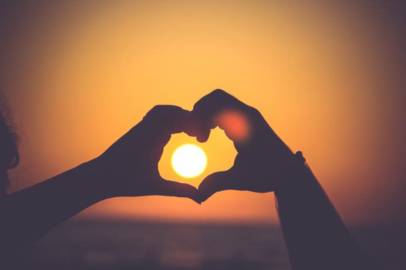 Two hands forming a heart shape against a dark violet sky with a soft golden glow between them.